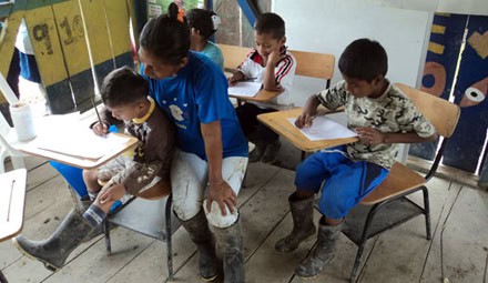 Teacher assisting young Colombian children writing in notebooks while seated at small desks in a classroom