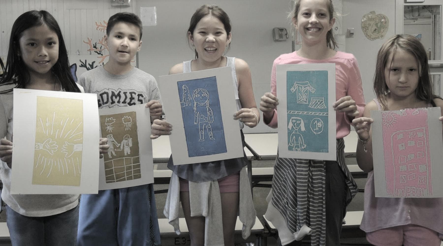 Five children standing in a classroom holding up their colorful art prints and smiling