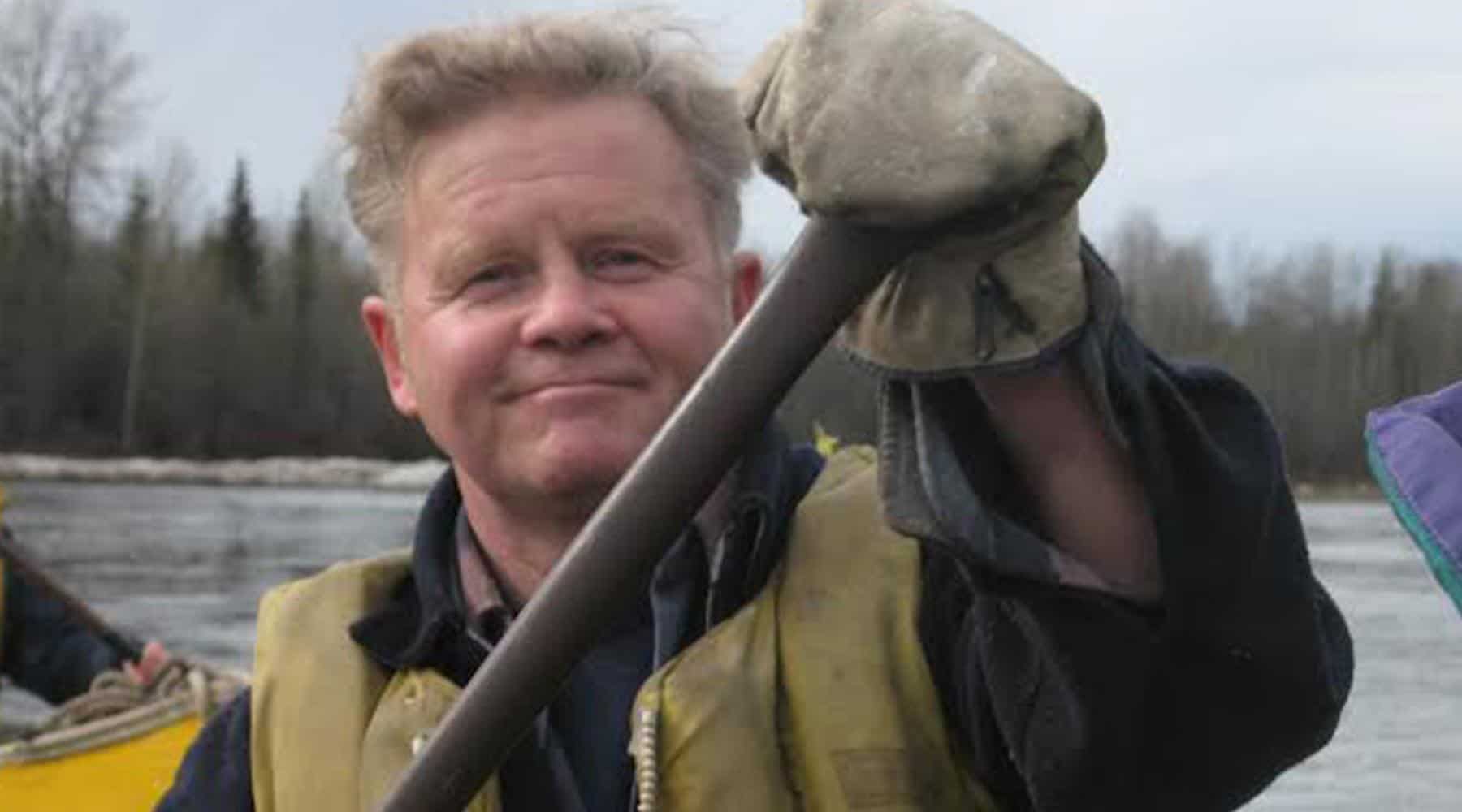 Peter Rodseth paddling a canoe on a river while wearing gloves and a life vest with forest in the background