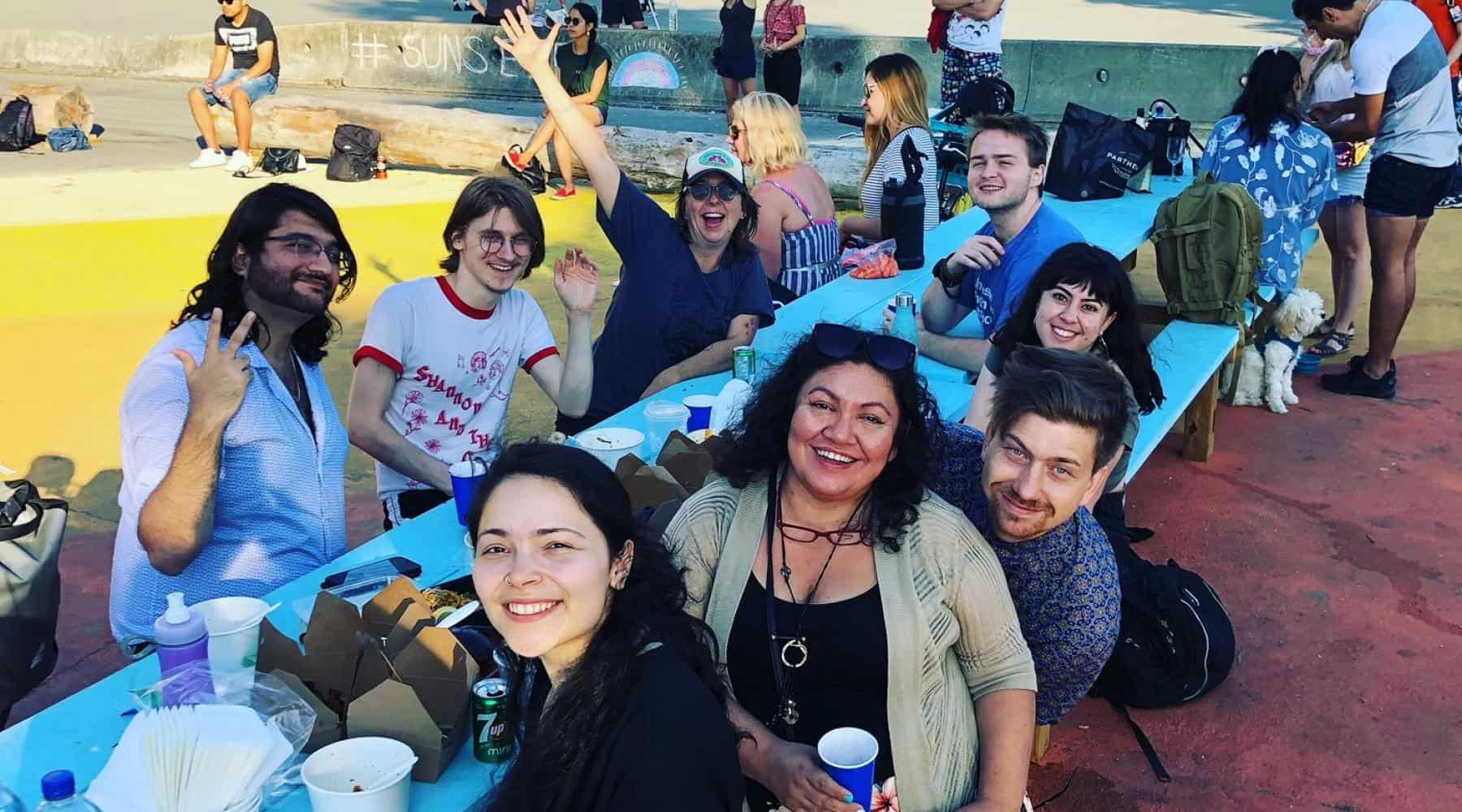 Group of people smiling and waving while sitting together at outdoor picnic tables during a community gathering.