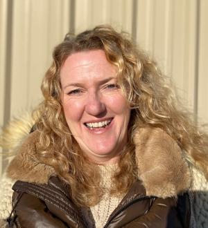 Heather O'Hara smiling, wearing a brown coat with a fur collar, standing outdoors in sunlight