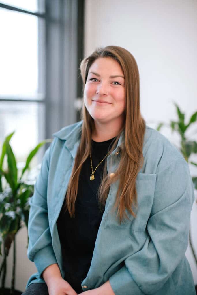 Smiling woman with long brown hair wearing a teal jacket seated in a bright office, with plants in the background
