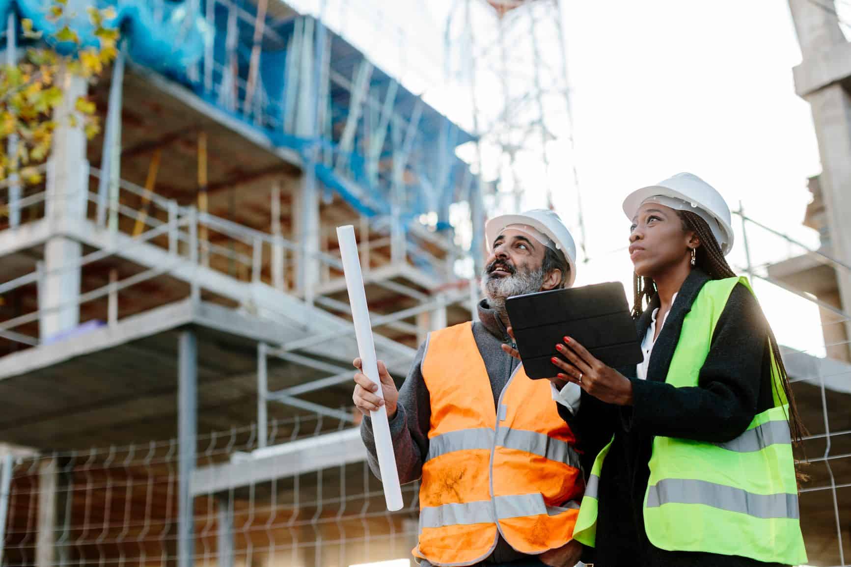 Two construction professionals wearing hard hats and safety vests reviewing plans at a building site