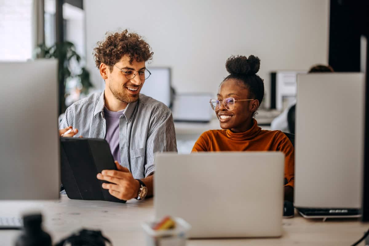 Two colleagues smiling and collaborating in front of computers in an office