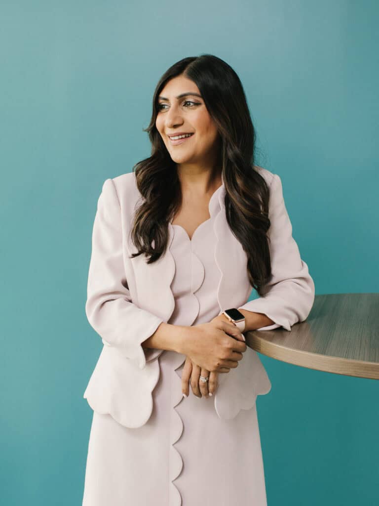 Professional portrait of a woman in a light blazer standing beside a table against a teal background.