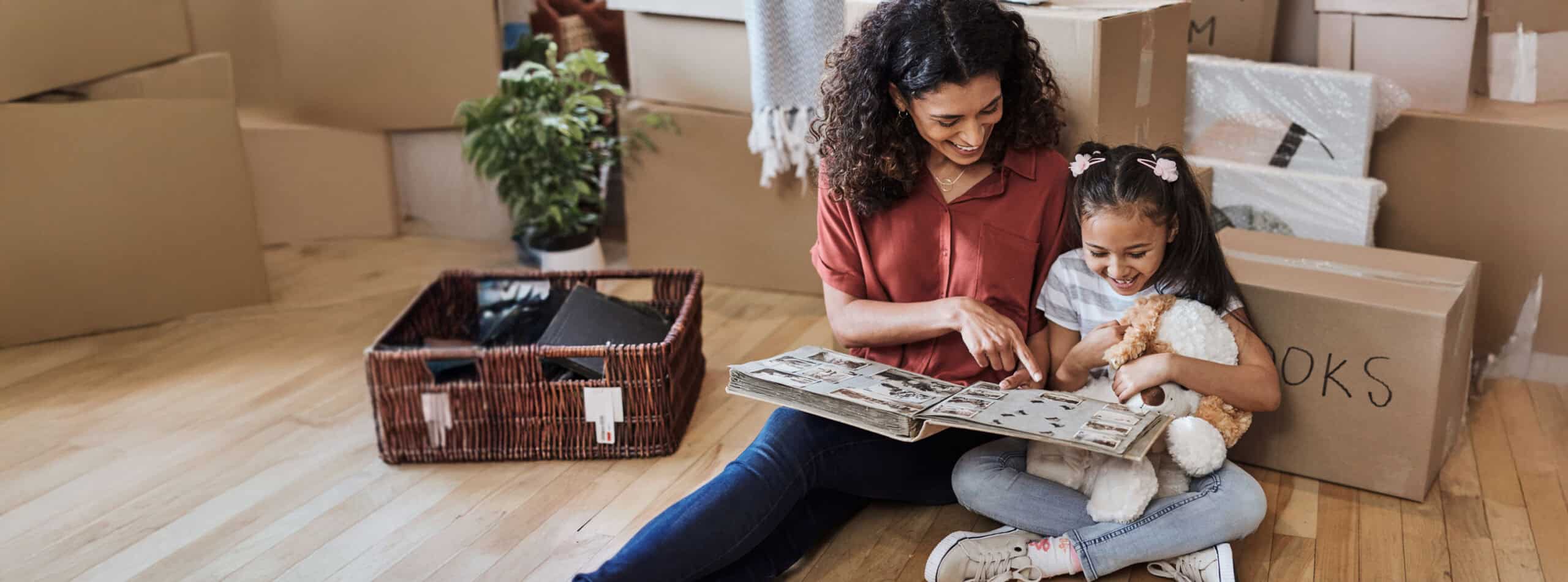 Mother and daughter sitting on the floor in a new home looking through a photo album surrounded by moving boxes.