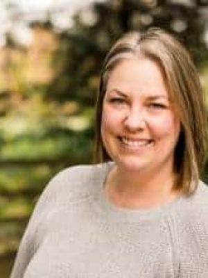 Woman smiling outdoors with trees and greenery in the background.