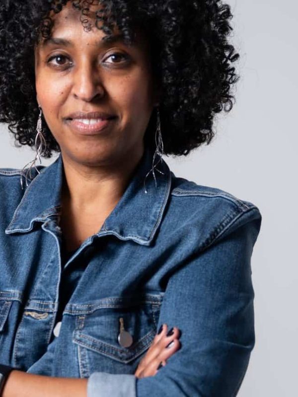 Woman with short curly hair wearing a denim jacket and earrings, standing with arms crossed against a neutral background