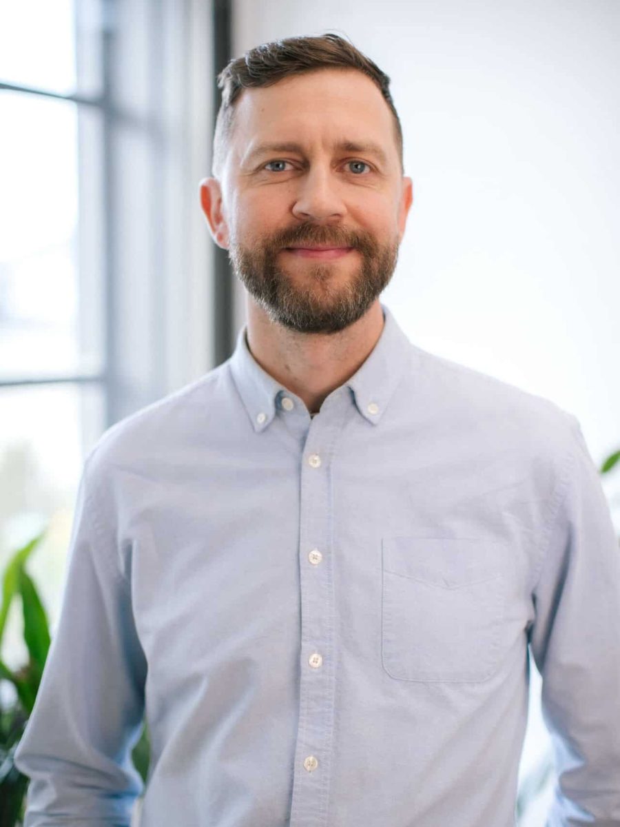 Smiling man with a beard wearing a light blue button-down shirt in a bright office