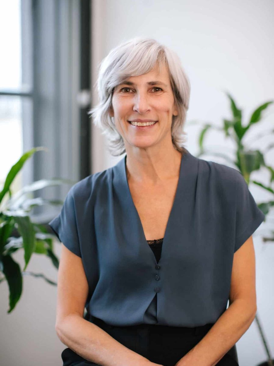 Smiling woman with short gray hair wearing a blue blouse seated in a bright office with plants in the background