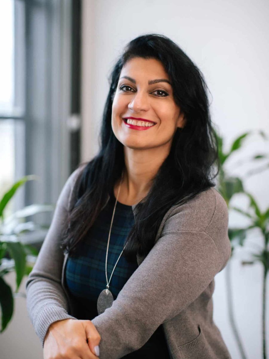 Smiling woman with long dark hair wearing a gray cardigan seated in a bright office, with plants in the background