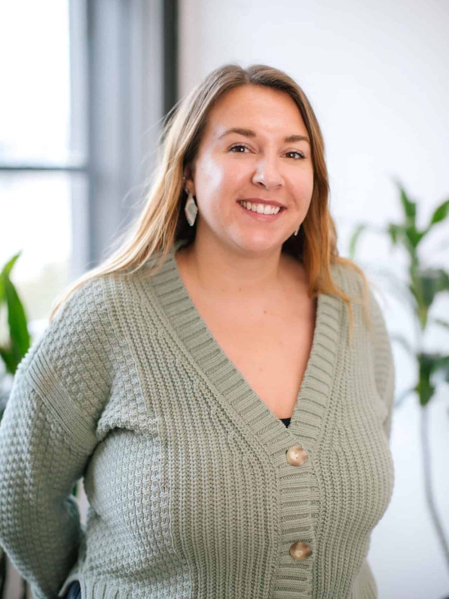 Smiling woman wearing an olive knit cardigan standing in a bright office with, plants in the background