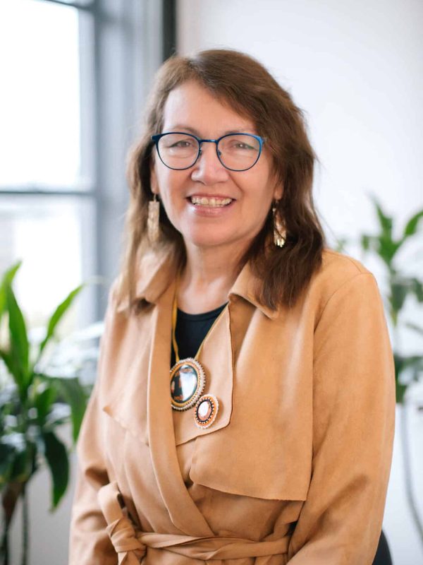 Smiling woman wearing blue glasses and a tan jacket seated in a bright office, with plants in the background