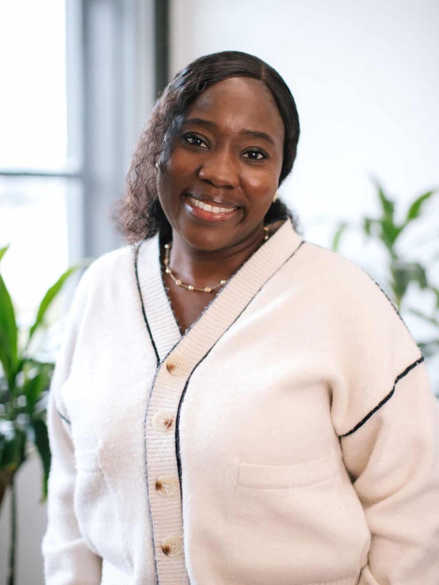 Smiling woman wearing a cream cardigan standing in a bright office with, plants in the background