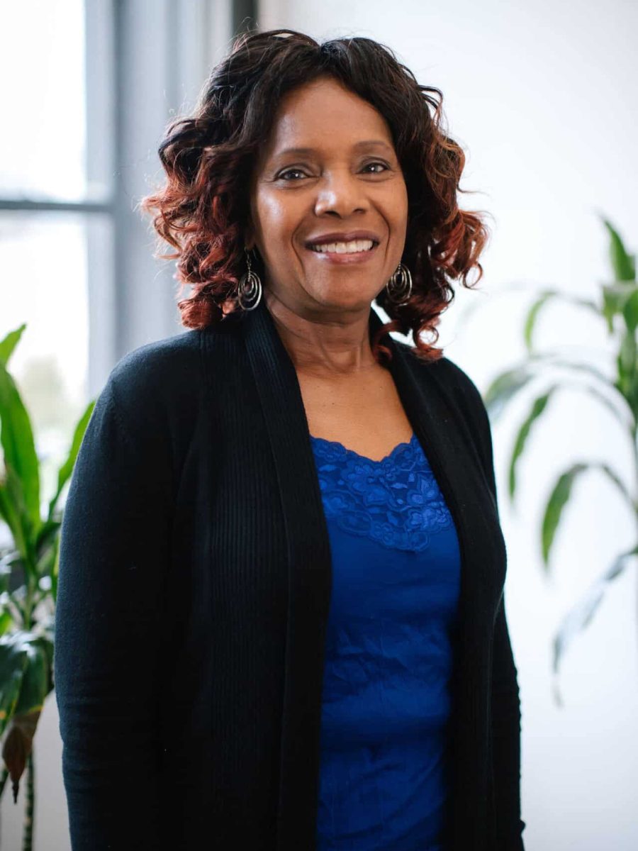 Smiling woman with curly hair wearing a blue top and black cardigan standing in a bright office with, plants in the background