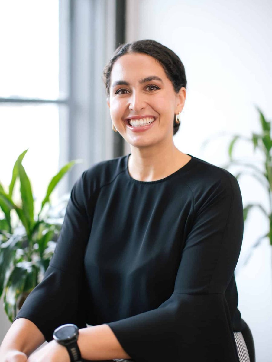Smiling woman with dark hair pulled back wearing a black top seated in a bright office, with plants in the background