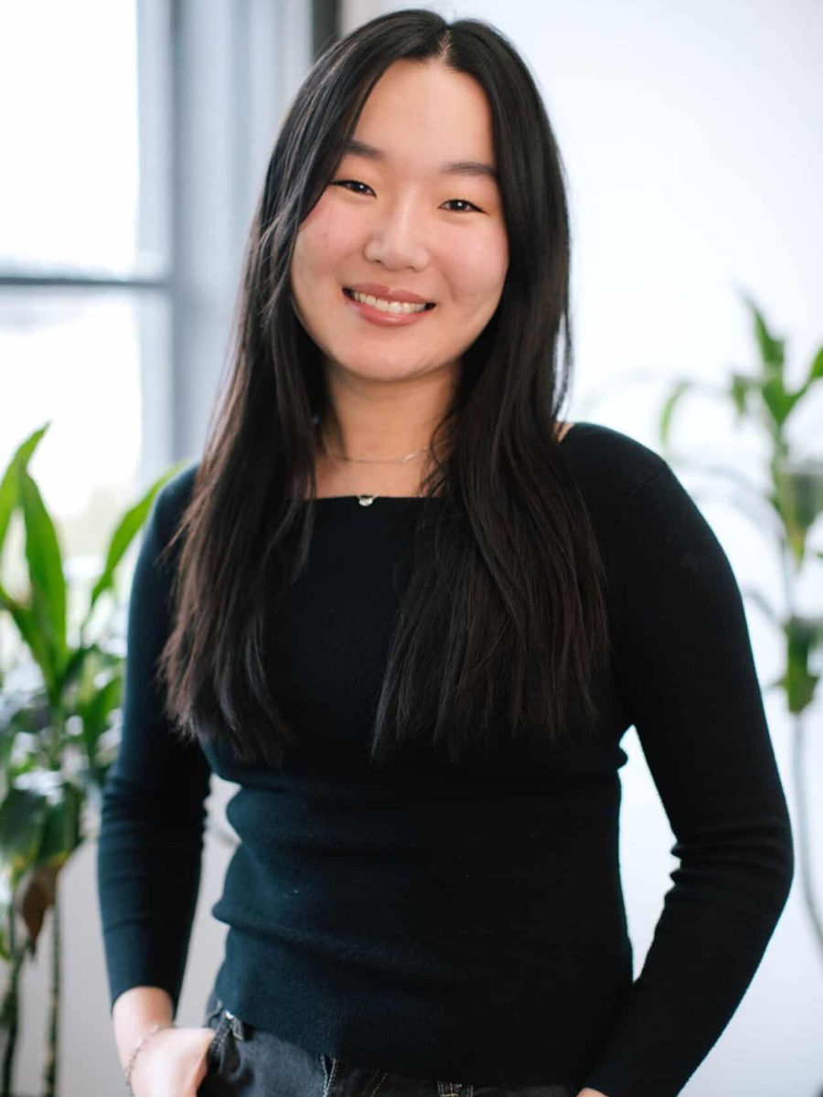 Smiling woman with long dark hair wearing a black top standing in a bright office, with plants in the background