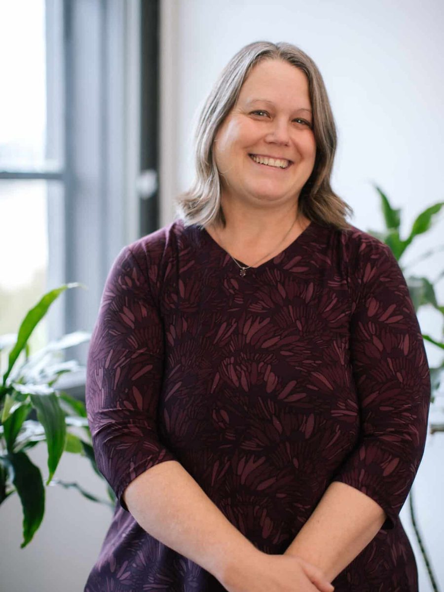 Smiling woman wearing a burgundy patterned dress standing in a bright office with plants in the background