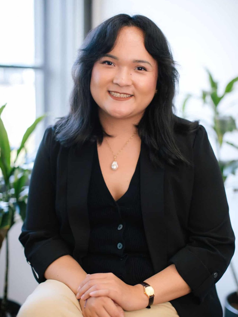 Smiling woman with shoulder-length dark hair wearing a black blazer seated in a bright office, with plants in the background