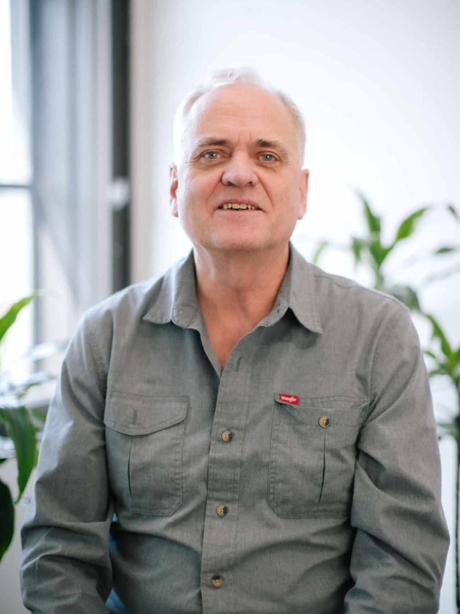 Smiling man wearing a gray button-up shirt seated in a bright office with plants, in the background