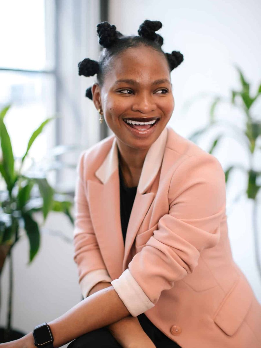 Smiling woman with bantu knot hairstyle wearing a blush blazer seated in a bright office, with plants in the background